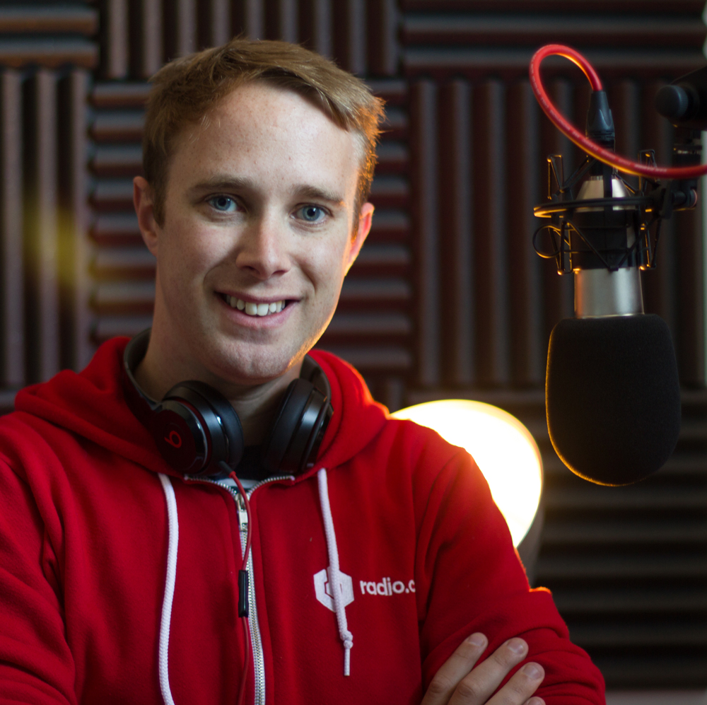This is an image of James Mulvany. He has brown hair and blue eyes, and smiles with his arms crossed at the camera. He is wearing a red Radio.co hoodie. The backdrop is a black studio with sound panels visible, and a black and red microphone in front of him.