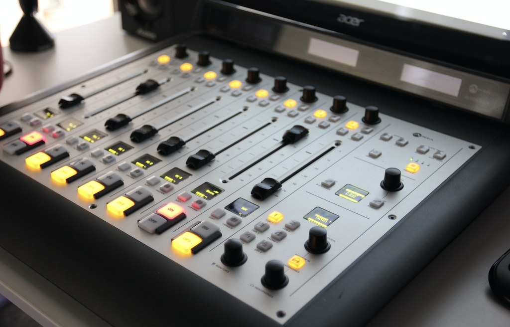 This is a silver Axia iQ Digital Mixing Desk with various lit up knobs and levels visible. It is is on a desk.