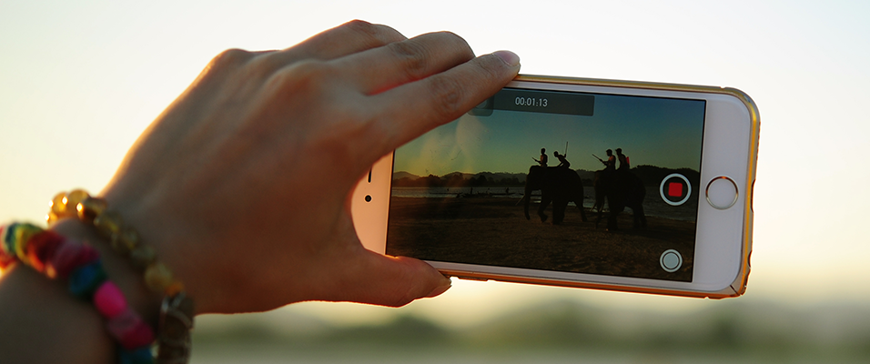 A white hand wearing bracelets holds up a white iPhone to a silhouettted group of people. We only see the image on the phone.