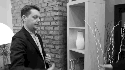 Shot in black and white, a white man with dark hair wearing a suit smiles and shakes hands with an offscreen arm.