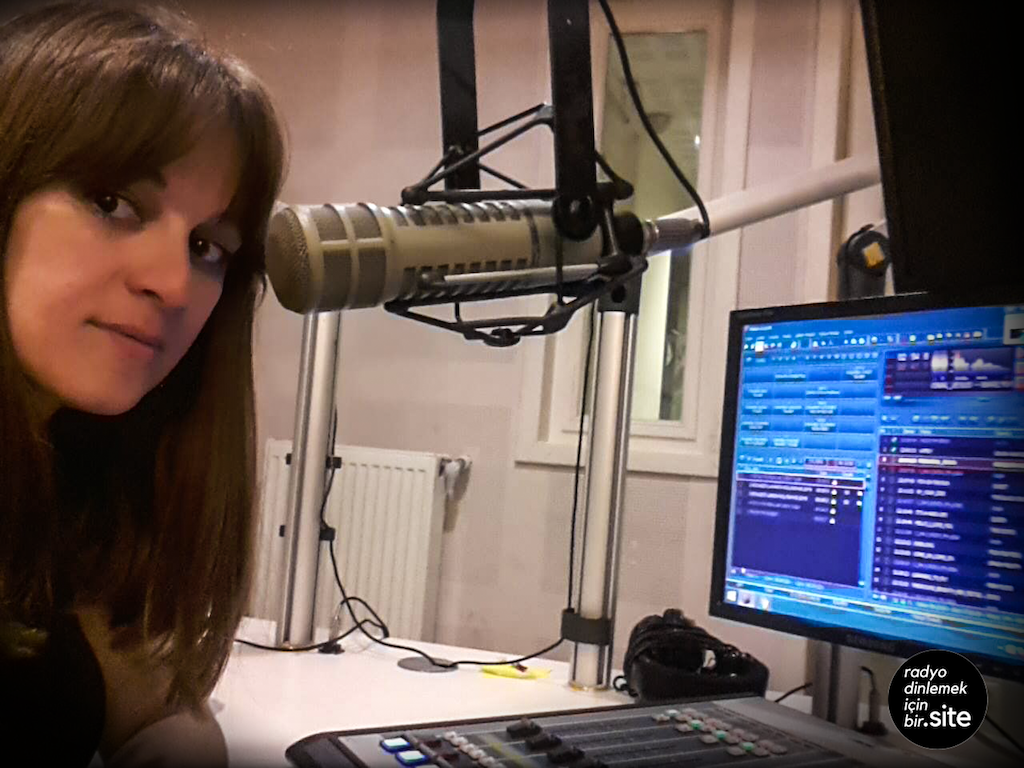 This is a white woman with brown eyes and long brunette hair. She is looking toward the camera and sits in front of a micrphone and a computer screen, additionally a mixing desk.