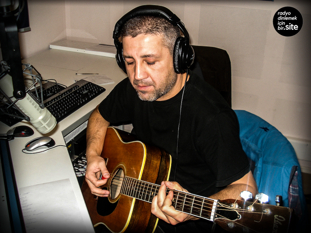This is a photo of a white man with grey hair and a grey short beard. He has his eyes shut and hsi playing a guitar in front of a microphone and mixing desk. He is wearing headphones. The RDiB Radio logo is in a black circle in the right hand corner