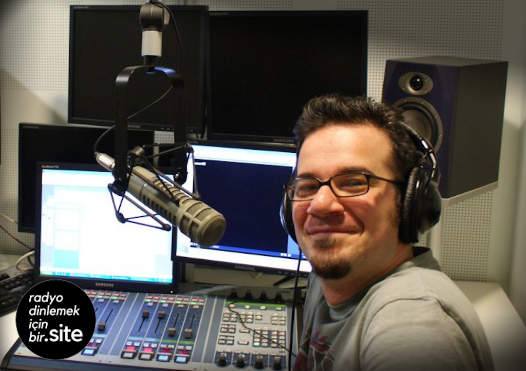 With the RDiB Radio logo in a black circle in the corner, this photo is of a white man with black hair and black glasses smiling at the camera. He is wearing headphones, smaller than the previous, and sitting in front of a micrphone and computer screen.