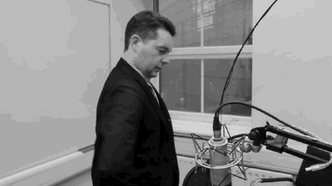 Shot in black and white, a white man with dark hair sits in front of a microphone and does reflex back stretches to warm himself up before speaking on the radio.