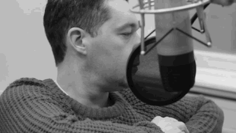 Shot in black and white, a white man with dark hair sits in front of a microphone and does reflex neck stretches to warm himself up before speaking on the radio.