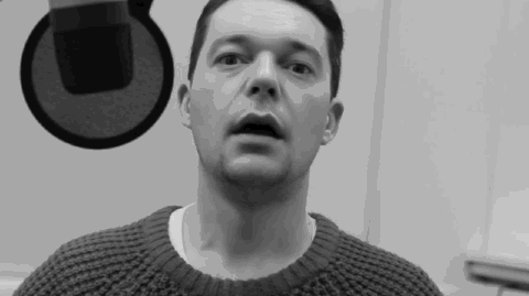 Shot in black and white, a white man with dark hair sits in front of a microphone and does reflex mouth stretches to warm himself up before speaking on the radio.